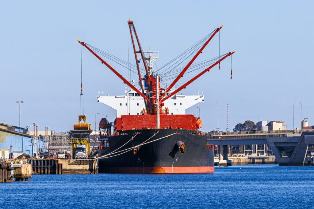 Large bulk carrier cargo ship unloading at port on a sunny day; symmetry with crane boom positions; large orange bucket (with "jaws" open) on crane boom loading/unloading large white semi-truck. Unrecognisable workers in hi-vis clothing on dock. Still, blue river water. Ship dominates skyline. ID, logos and flag edited, tight crop